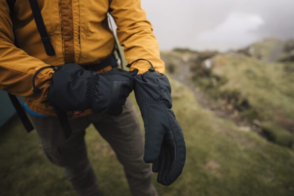 Defence-gloves-5.jpg Person in a mustard-yellow jacket and brown trousers fastening black insulated gloves while standing on a misty grassy hillside.