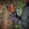 Drax-Glove-scaled-4.jpg Person in a dark green jacket pulling on insulated black gloves with brown leather palms while standing beside a tree trunk in a blurred woodland background.