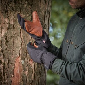 Person in a dark green jacket pulling on insulated black gloves with brown leather palms while standing beside a tree trunk in a blurred woodland background.