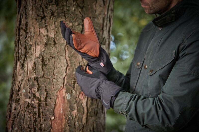 Drax-Glove-scaled-4.jpg Person in a dark green jacket pulling on insulated black gloves with brown leather palms while standing beside a tree trunk in a blurred woodland background.