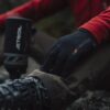 EXTREMITIES_PHOTOGRAPHY_2023_0026-3.jpg Close-up of a hiker's gloved hand wearing a black extremities glove resting on a muddy trouser knee, red jacket sleeve visible, with a hiking boot and blurred Jetboil stove nearby.