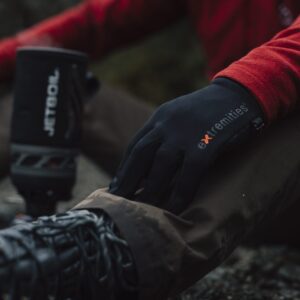 Close-up of a hiker's gloved hand wearing a black extremities glove resting on a muddy trouser knee, red jacket sleeve visible, with a hiking boot and blurred Jetboil stove nearby.