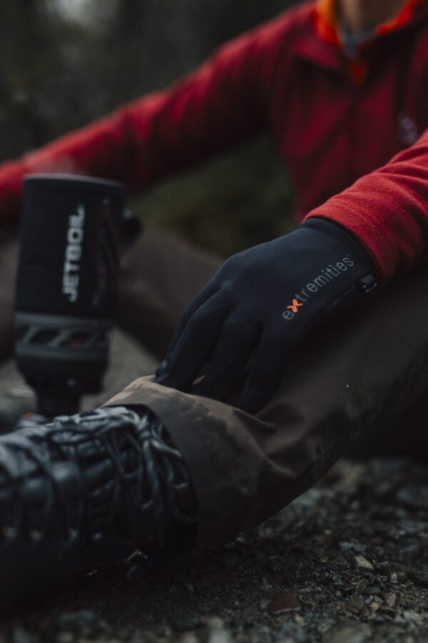 EXTREMITIES_PHOTOGRAPHY_2023_0026-3.jpg Close-up of a hiker's gloved hand wearing a black extremities glove resting on a muddy trouser knee, red jacket sleeve visible, with a hiking boot and blurred Jetboil stove nearby.