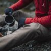 EXTREMITIES_PHOTOGRAPHY_2023_0031-3.jpg Person in a red fleece and black gloves sitting on a rocky riverbank, pouring water from a camping pot into a metal cup beside a small portable gas stove.