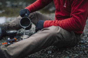 EXTREMITIES_PHOTOGRAPHY_2023_0031-3.jpg Person in a red fleece and black gloves sitting on a rocky riverbank, pouring water from a camping pot into a metal cup beside a small portable gas stove.