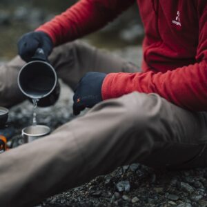 Person in a red fleece and black gloves sitting on a rocky riverbank, pouring water from a camping pot into a metal cup beside a small portable gas stove.