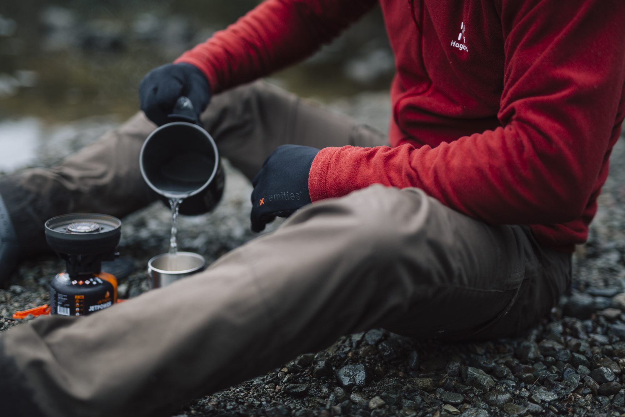 EXTREMITIES_PHOTOGRAPHY_2023_0031-3.jpg Person in a red fleece and black gloves sitting on a rocky riverbank, pouring water from a camping pot into a metal cup beside a small portable gas stove.
