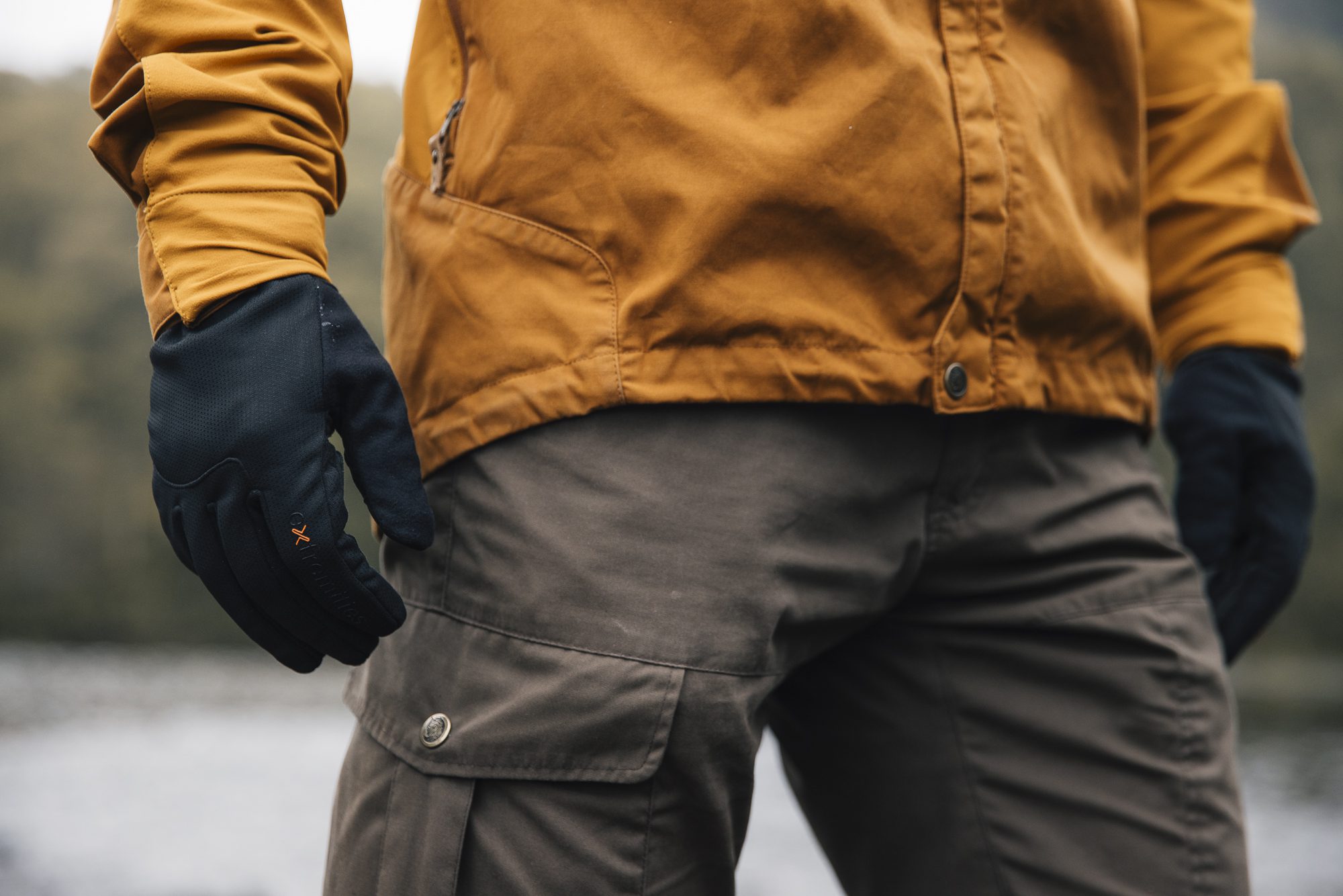 Close-up of a person's lower torso and hands standing by water, wearing a mustard-yellow jacket, black gloves with a small orange logo and grey cargo trousers