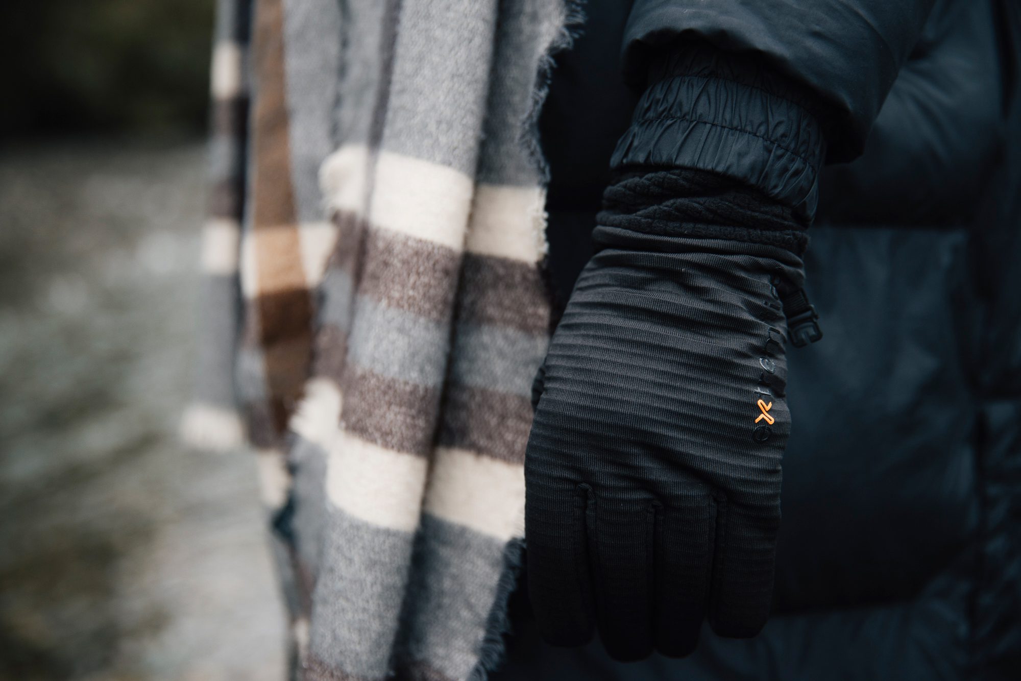 Close-up of a person outdoors near water wearing a black insulated glove with a small orange logo and a dark puffer sleeve, beside a grey, brown and cream striped wool scarf.