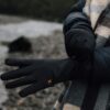 Person on a stony shoreline wearing a dark puffer jacket and checked scarf adjusts a pair of black insulated gloves with a small orange logo.