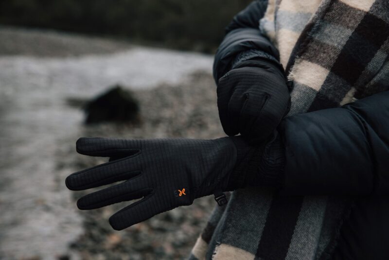EXTREMITIES_PHOTOGRAPHY_2023_0084-3.jpg Person on a stony shoreline wearing a dark puffer jacket and checked scarf adjusts a pair of black insulated gloves with a small orange logo.