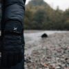 Person's arm in a black puffer coat wearing a black quilted Extremities glove next to a grey-and-cream striped scarf, standing on a pebble riverbank with trees blurred in the background.