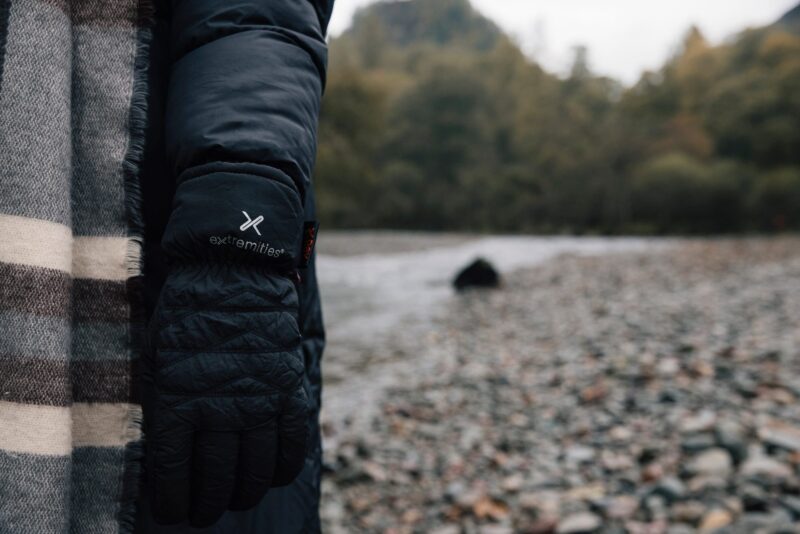 EXTREMITIES_PHOTOGRAPHY_2023_0092-3.jpg Person's arm in a black puffer coat wearing a black quilted Extremities glove next to a grey-and-cream striped scarf, standing on a pebble riverbank with trees blurred in the background.
