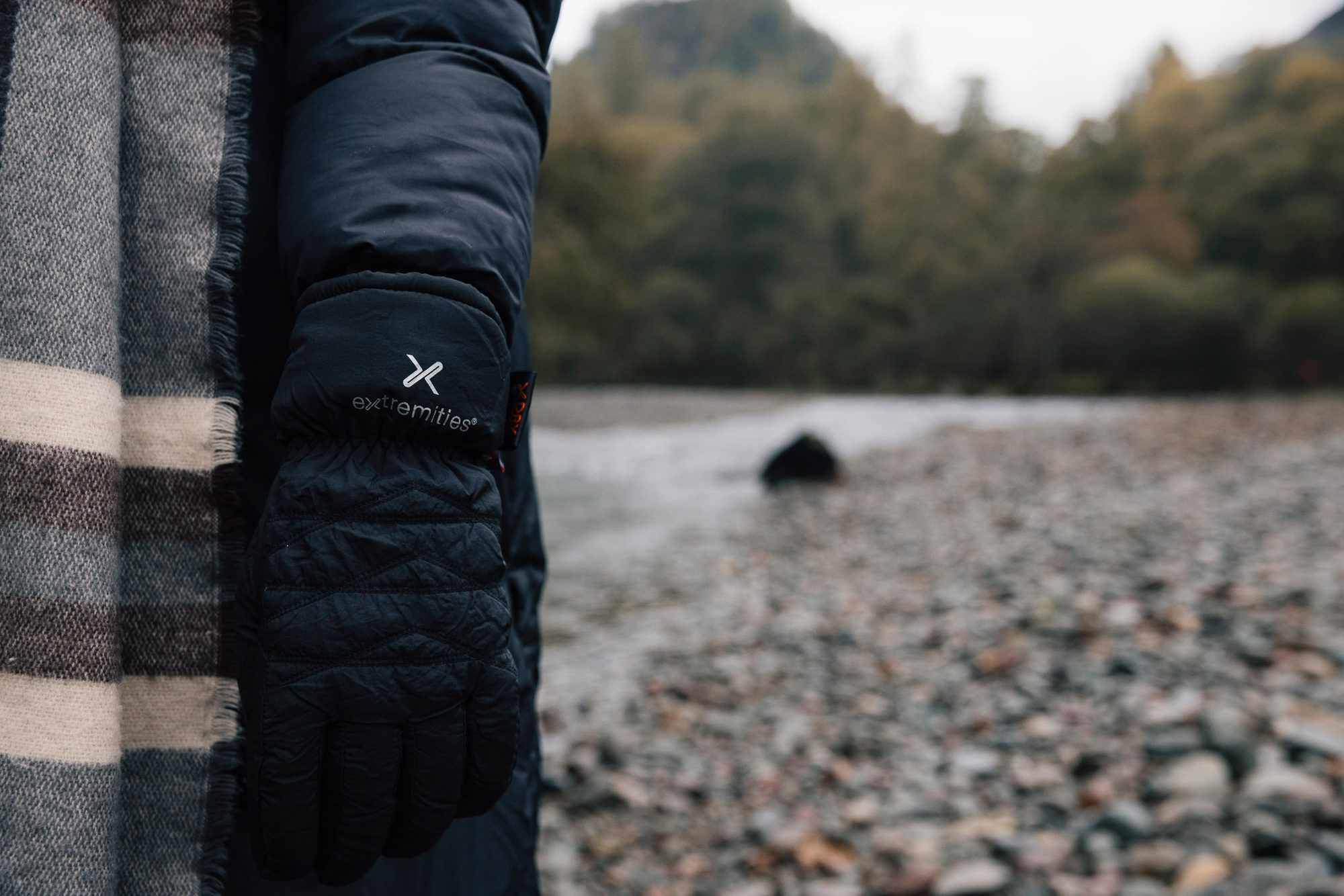 Person's arm in a black puffer coat wearing a black quilted Extremities glove next to a grey-and-cream striped scarf, standing on a pebble riverbank with trees blurred in the background.