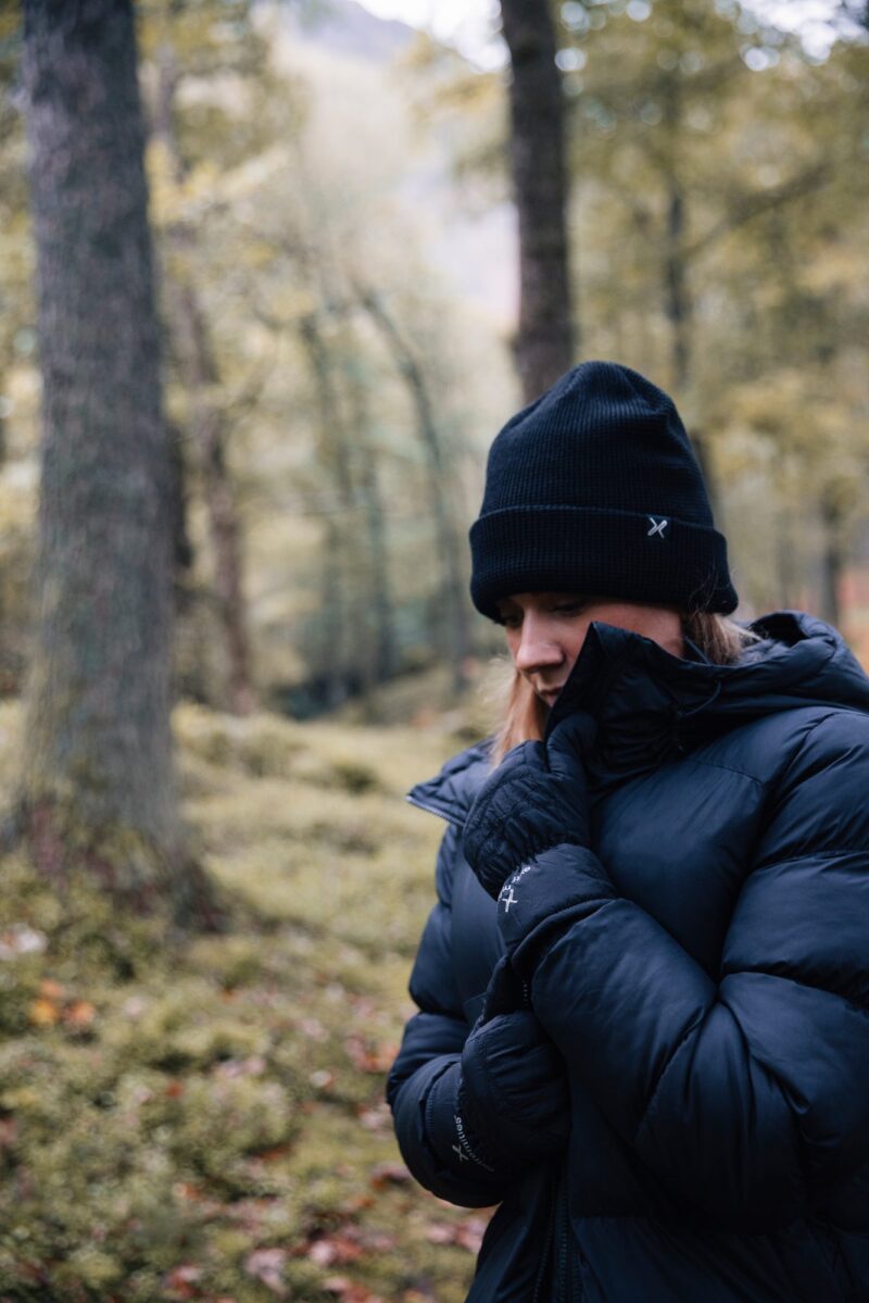 EXTREMITIES_PHOTOGRAPHY_2023_0113-3.jpg A person in a black beanie, dark padded winter jacket and gloves pulls the collar up around their face and looks down while standing in a mossy, tree-filled forest.