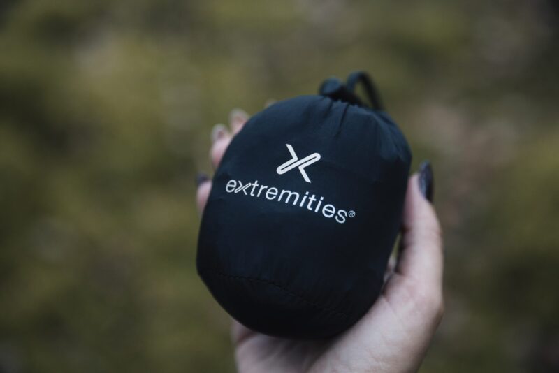 EXTREMITIES_PHOTOGRAPHY_2023_0126-3.jpg Hand holding a small black drawstring stuff sack stamped with a white "extremities" logo against a blurred green outdoor background.