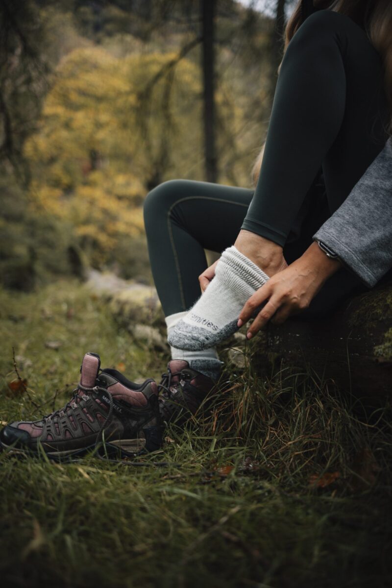 EXTREMITIES_PHOTOGRAPHY_2023_0142-3.jpg Person sitting on a mossy log in an autumn woodland, pulling a thick grey hiking sock onto one foot while maroon hiking boots rest on the grass beside them.