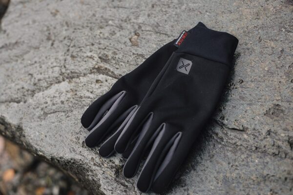 EXTREMITIES_PHOTOGRAPHY_2023_0143-3.jpg Black lightweight gloves with grey fingertip panels and a small logo, laid flat on a rough grey stone surface