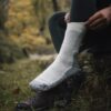 EXTREMITIES_PHOTOGRAPHY_2023_0144-3.jpg Close-up of a person pulling a thick white wool hiking sock over dark green leggings with their foot resting on a brown hiking shoe in a grassy, autumnal forest setting.