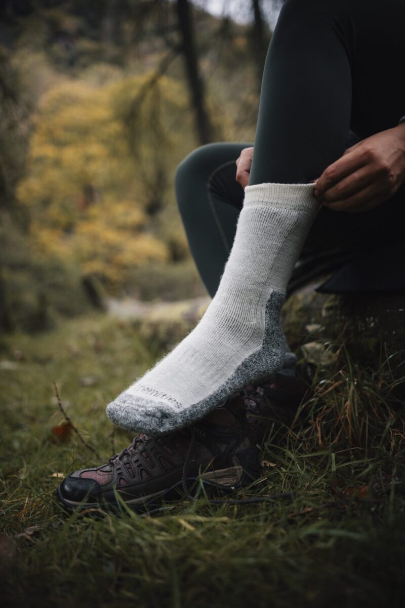 EXTREMITIES_PHOTOGRAPHY_2023_0144-3.jpg Close-up of a person pulling a thick white wool hiking sock over dark green leggings with their foot resting on a brown hiking shoe in a grassy, autumnal forest setting.