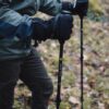 EXTREMITIES_PHOTOGRAPHY_2023_0233-3.jpg Close-up of a hiker's hands in black insulated gloves gripping Terra Nova carbon trekking poles, wearing a green jacket and brown trousers while standing on a mossy, leaf-strewn forest floor.