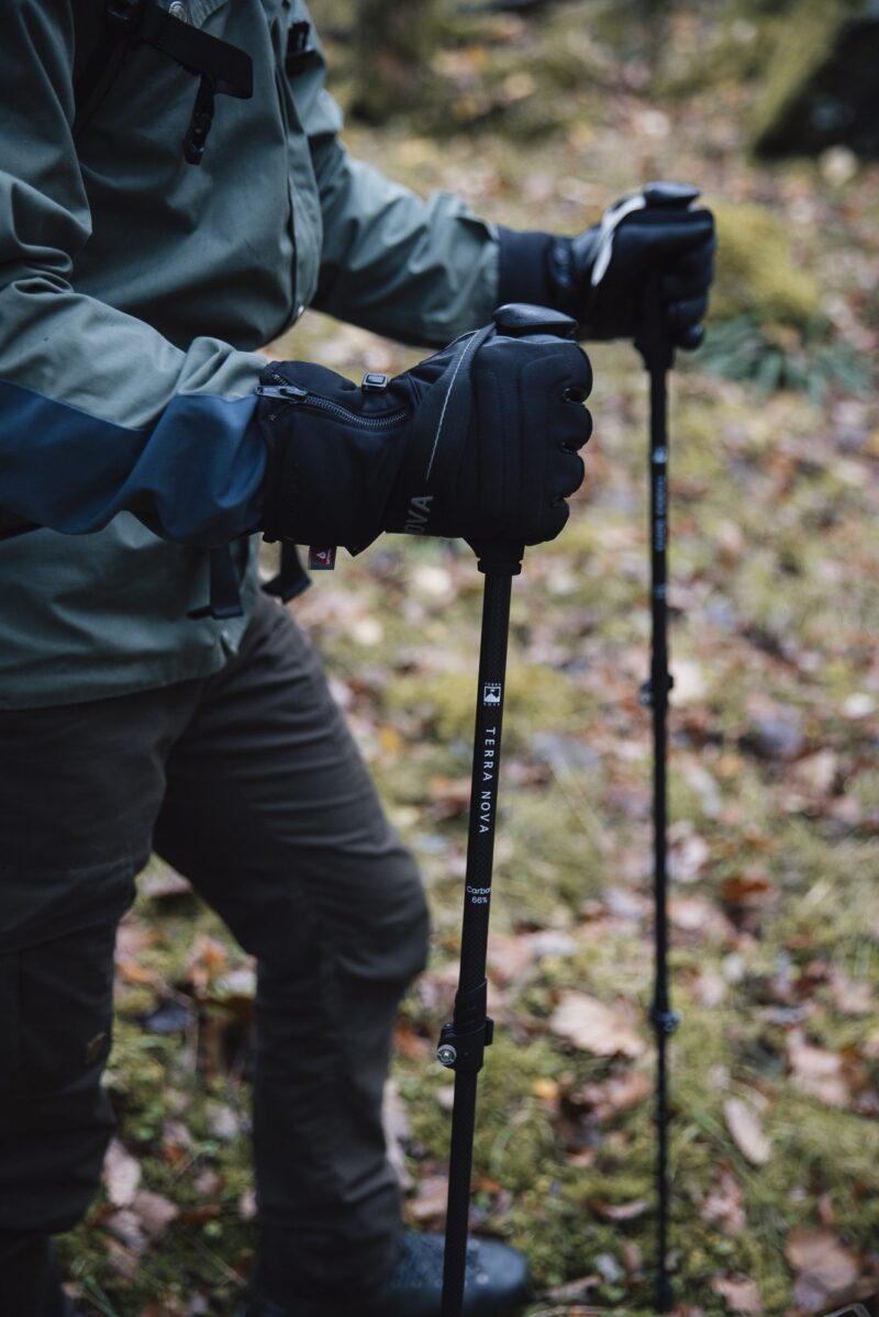 Close-up of a hiker's hands in black insulated gloves gripping Terra Nova carbon trekking poles, wearing a green jacket and brown trousers while standing on a mossy, leaf-strewn forest floor.