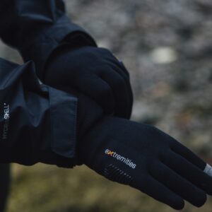 Close-up of a person’s crossed hands wearing black Extremities touchscreen gloves with a grey fingertip and a black Hydroshell jacket sleeve against a blurred outdoor ground.