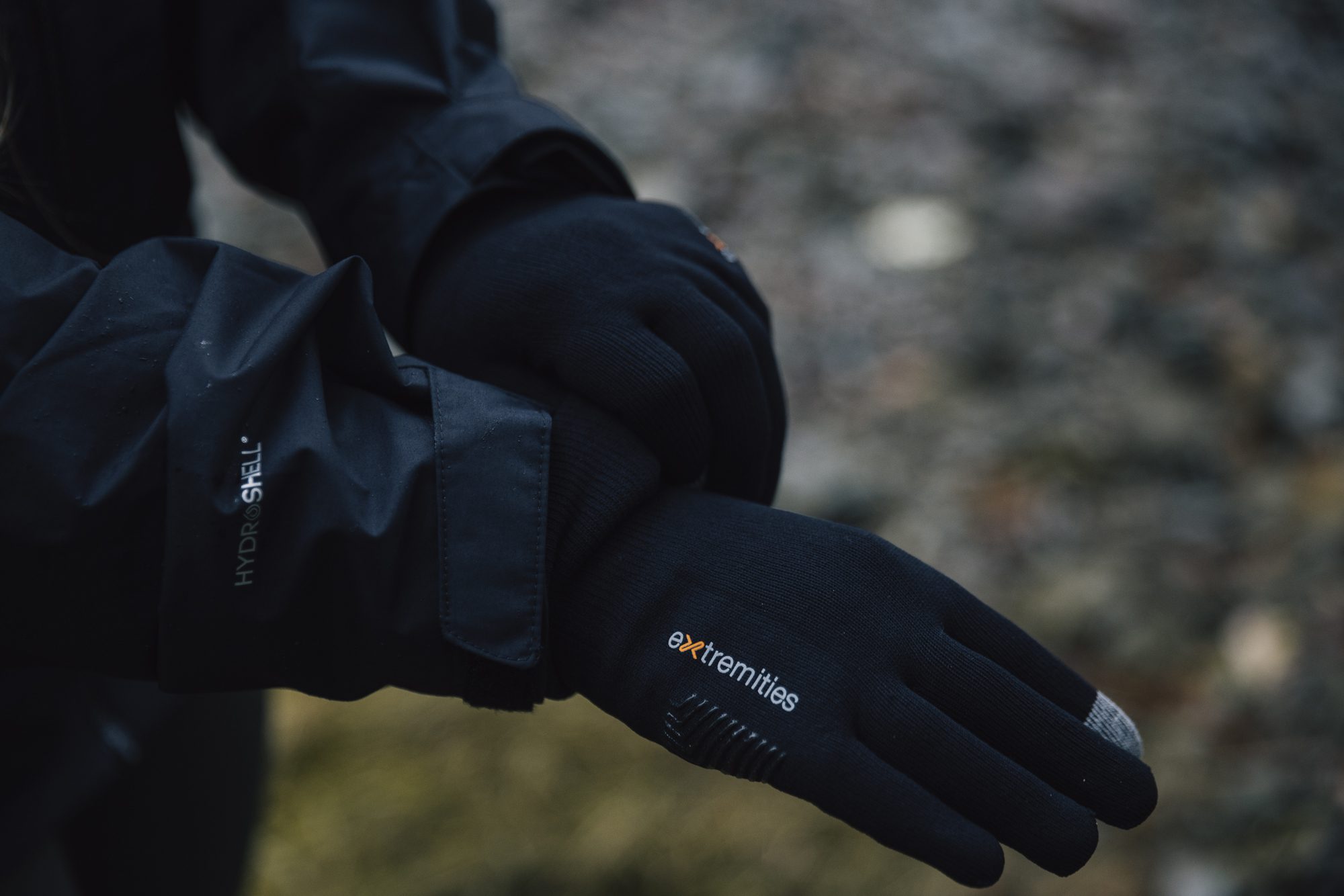 Close-up of a person’s crossed hands wearing black Extremities touchscreen gloves with a grey fingertip and a black Hydroshell jacket sleeve against a blurred outdoor ground.