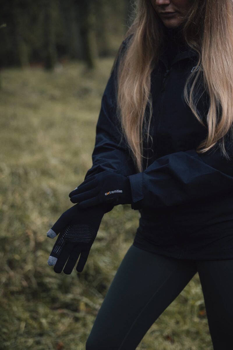 Person in a black jacket and green leggings stands in a grassy woodland, adjusting black touchscreen gloves with grey fingertips labelled extremities, long blonde hair falling over their shoulders.