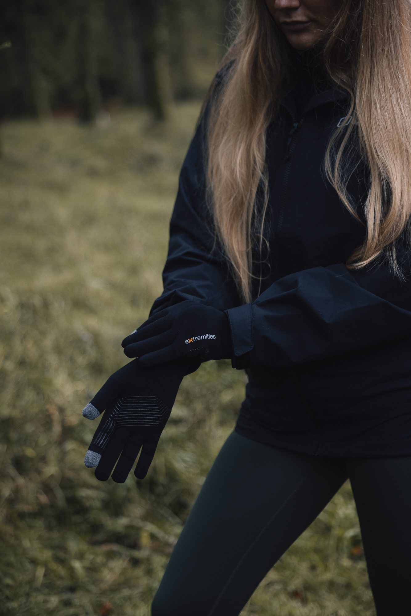 Person in a black jacket and green leggings stands in a grassy woodland, adjusting black touchscreen gloves with grey fingertips labelled extremities, long blonde hair falling over their shoulders.