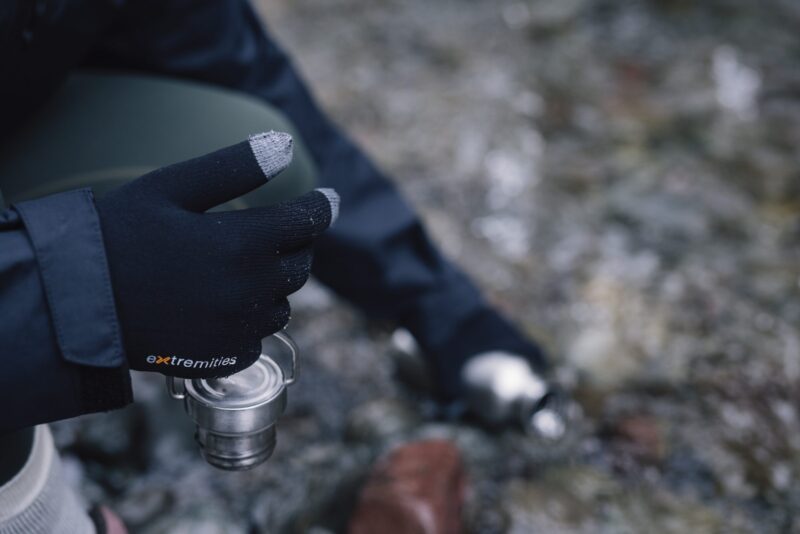 Close-up of a gloved hand wearing black Extremities touchscreen gloves with grey fingertips holding a small stainless-steel camping cup above a rocky, out-of-focus ground with a metal water bottle blurred in the background.