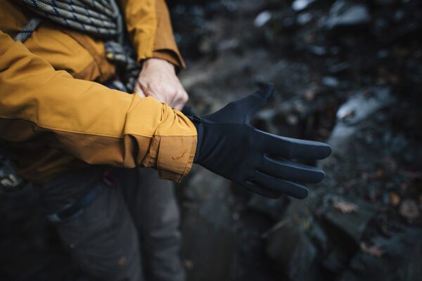 EXTREMITIES_PHOTOGRAPHY_2023_0362-3.jpg Person outdoors pulling a black glove onto their hand over a mustard-yellow jacket sleeve, rocky ground visible in the background.