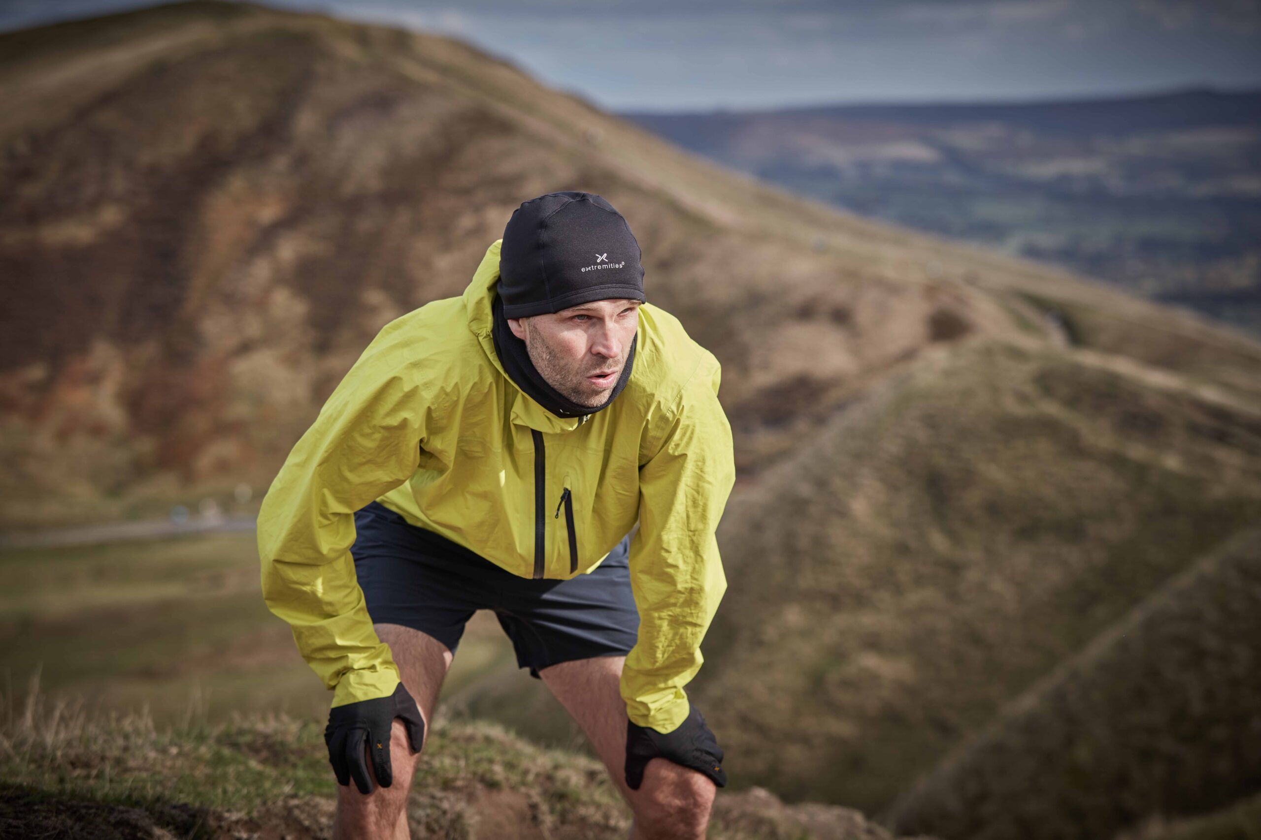 Man in a bright yellow windproof jacket, black beanie and gloves leaning forward with hands on his knees, panting during a pause on a grassy hill with rolling moorland behind him.