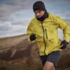Runner wearing a bright yellow waterproof jacket, black beanie, neck gaiter, gloves and grey shorts sprinting across windswept hills under an overcast sky.