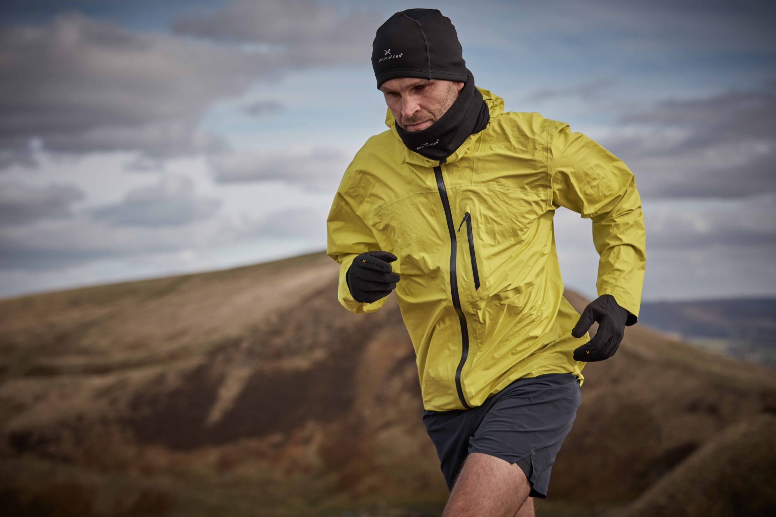 Runner wearing a bright yellow waterproof jacket, black beanie, neck gaiter, gloves and grey shorts sprinting across windswept hills under an overcast sky.