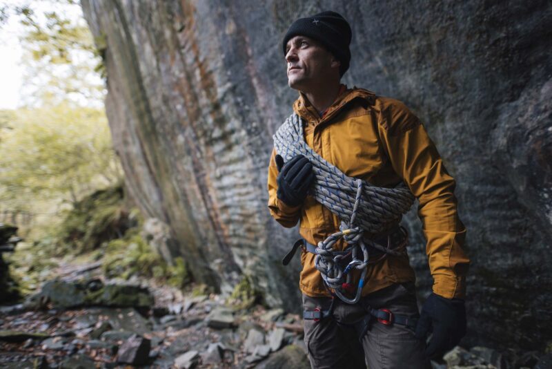 Climber in a mustard jacket and black beanie stands beside a steep rock face with a coiled rope over his shoulder and a harness with carabiners, looking thoughtfully to the right amid mossy rocks and trees.