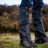 Edited-Image-NEW-ORANGE-2-3.jpg Close-up of lower legs wearing black waterproof gaiters over hiking boots and blue jeans, water droplets beading on the fabric as the wearer stands on grassy, muddy ground with blurred heathland behind,