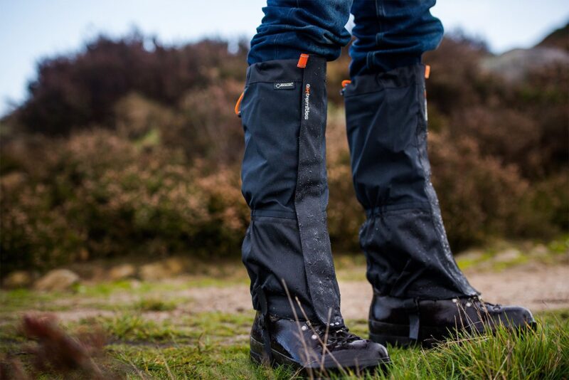 Edited-Image-NEW-ORANGE-2-3.jpg Close-up of lower legs wearing black waterproof gaiters over hiking boots and blue jeans, water droplets beading on the fabric as the wearer stands on grassy, muddy ground with blurred heathland behind,