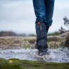 Edited-Image-NEW-ORANGE-4.jpg Lower legs in blue jeans and black waterproof gaiters and boots splashing through a muddy puddle on a windswept moorland path, water droplets frozen mid‑air against an overcast sky.