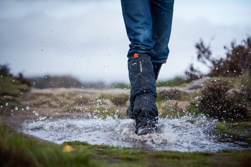Edited-Image-NEW-ORANGE-4.jpg Lower legs in blue jeans and black waterproof gaiters and boots splashing through a muddy puddle on a windswept moorland path, water droplets frozen mid‑air against an overcast sky.