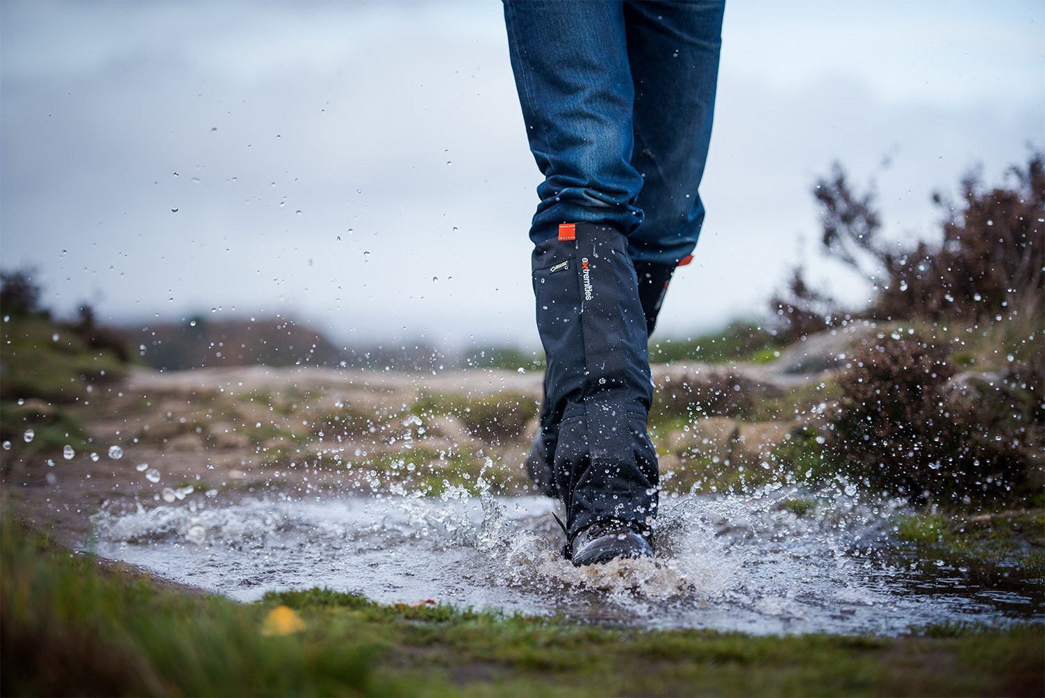 Lower legs in blue jeans and black waterproof gaiters and boots splashing through a muddy puddle on a windswept moorland path, water droplets frozen mid‑air against an overcast sky.