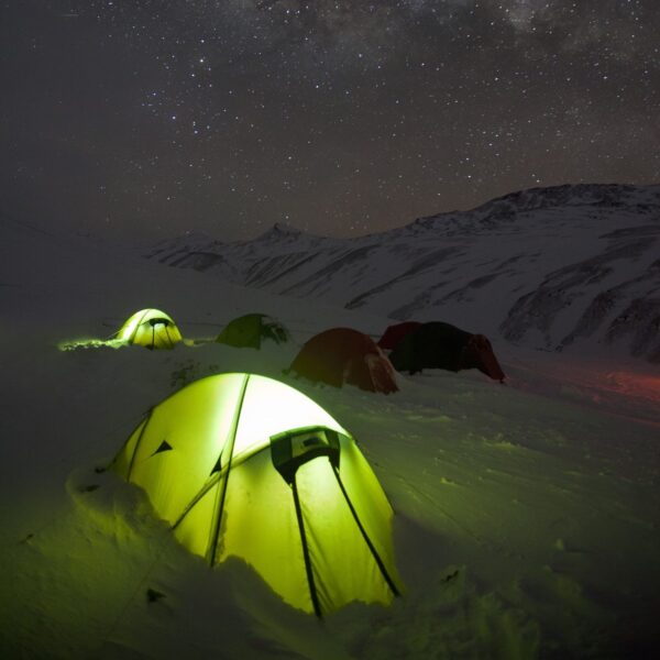 Expedition-Quasar-Lifestyle-3.jpg Glowing yellow-green tent in the snowy foreground with several other tents pitched on a snow-covered mountain slope beneath a clear, star-filled night sky and distant peaks.