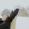 Extremities-Japan-Low-Res-15-scaled-4.jpg Person wearing a grey beanie and black gloves presses gloved hands against the tip of a patterned ski in a snowy landscape with snow-covered trees in the background.