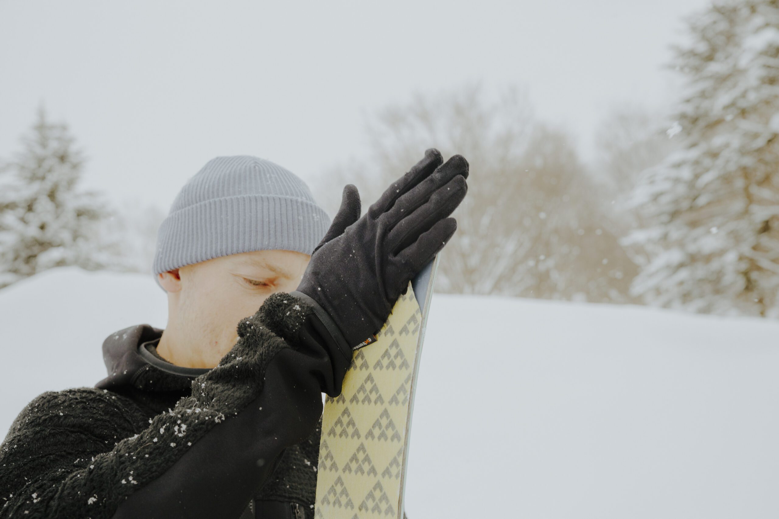 Person wearing a grey beanie and black gloves presses gloved hands against the tip of a patterned ski in a snowy landscape with snow-covered trees in the background.