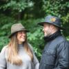 Extremities-shoot-15.8-4-scaled-2.jpg A smiling woman in a grey ribbed sweater and brown fedora talks to a bearded man in a black jacket and wide-brimmed hat decorated with a colourful feather, standing together in a leafy outdoor setting.
