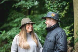 A smiling woman in a grey ribbed sweater and brown fedora talks to a bearded man in a black jacket and wide-brimmed hat decorated with a colourful feather, standing together in a leafy outdoor setting.