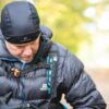 Extremitieshoot_Oct2019-110-scaled-2.jpg Middle-aged man wearing a black beanie and quilted down jacket with backpack straps and blue trim, looking down while standing on a blurred autumn woodland path.