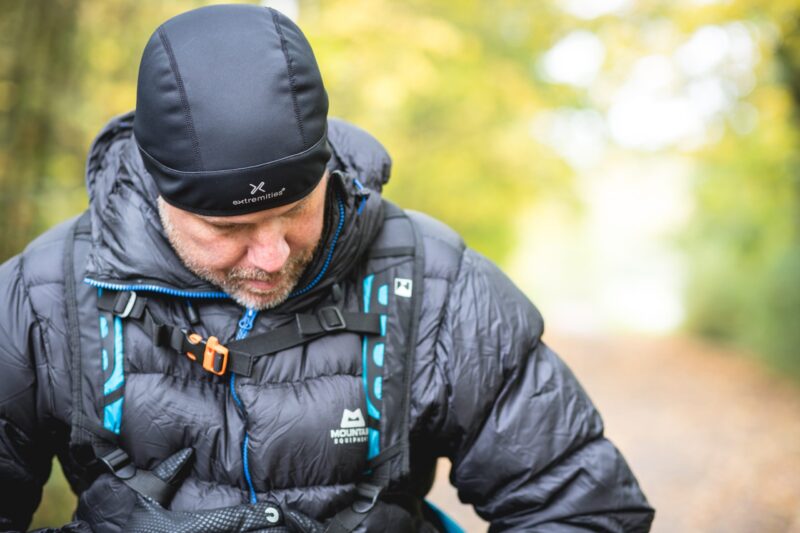 Extremitieshoot_Oct2019-110-scaled-2.jpg Middle-aged man wearing a black beanie and quilted down jacket with backpack straps and blue trim, looking down while standing on a blurred autumn woodland path.