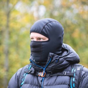 Person wearing a black balaclava and puffy dark jacket with blue-accented backpack straps, looking to the left against a blurred autumnal woodland background.