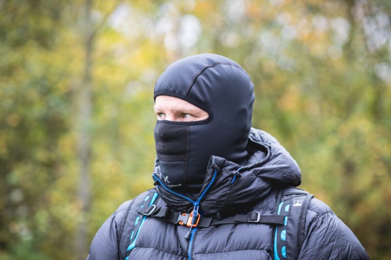 Person wearing a black balaclava and puffy dark jacket with blue-accented backpack straps, looking to the left against a blurred autumnal woodland background.